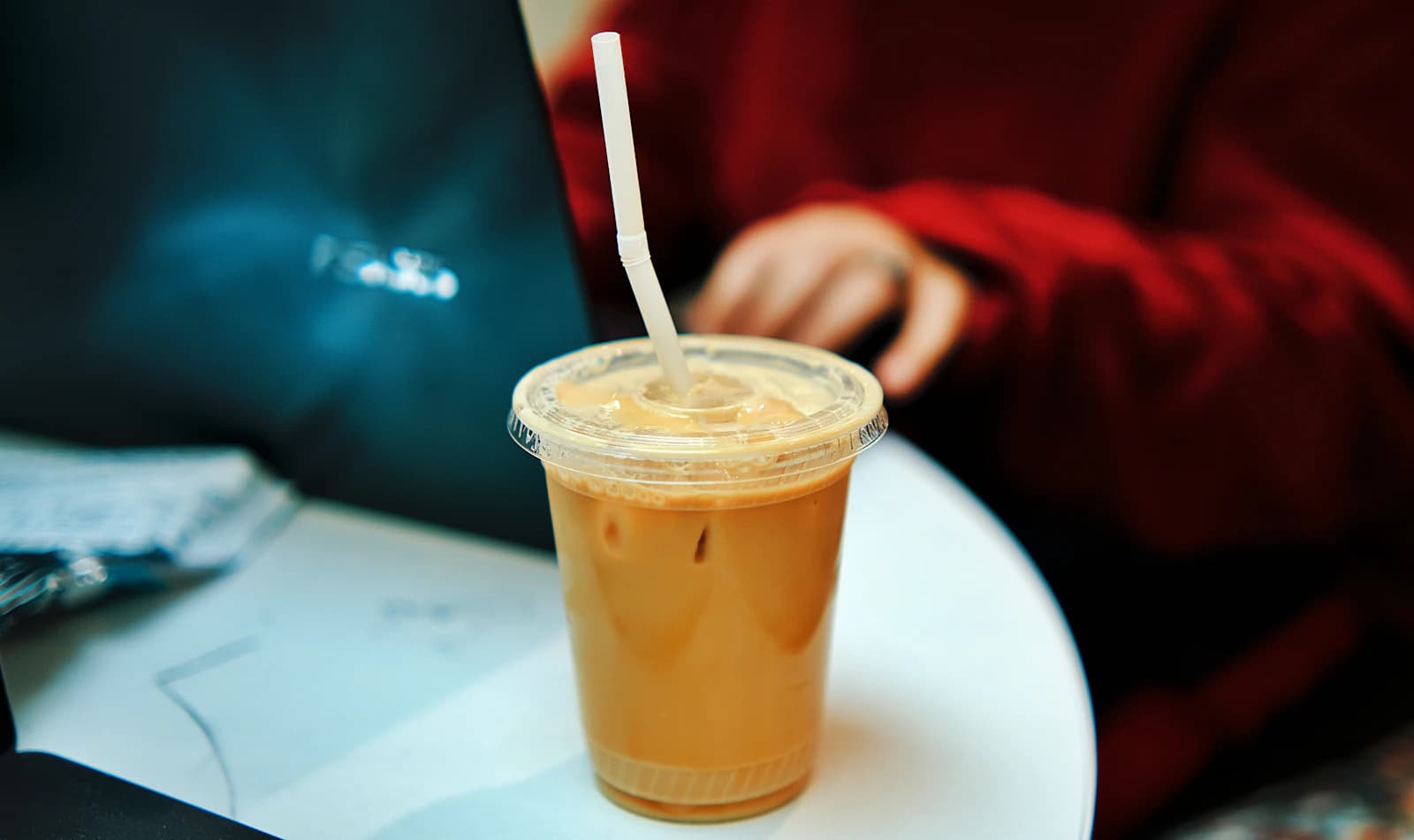 An iced latte in a takeaway cup with a straw on a table beside a laptop.