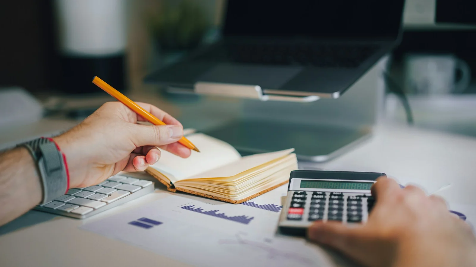 A person calculating costs with a calculator, notebook and financial charts.