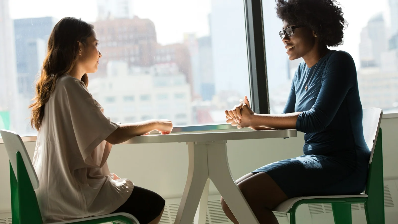 Two colleagues having a conversation at a table beside an office window.